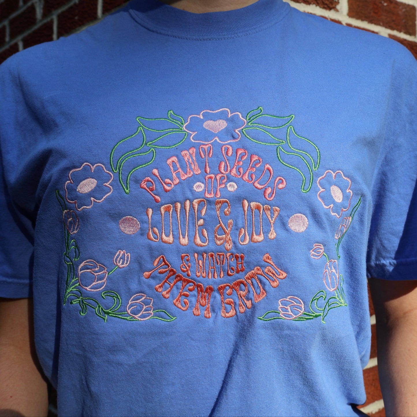 Blue t-shirt with floral design and text against a brick wall background